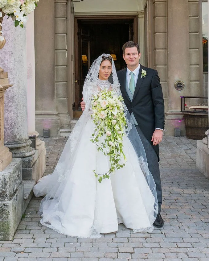 A bride and groom standing together in front of a building entrance on their wedding day. The bride is wearing a white wedding gown with lace details and a veil, holding a cascading bouquet of white and green flowers. The groom is dressed in a dark suit with a white shirt and light blue tie, with a boutonniere on his lapel.
