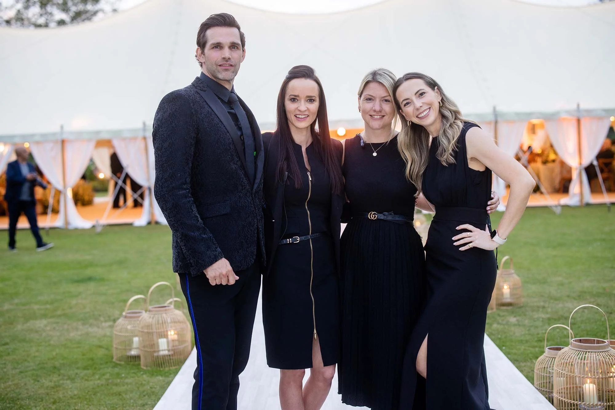 Four people, two men and two women, dressed in formal black attire, standing outdoors at an event with a white tent and lanterns in the background. Cranberry Blue's team at work.