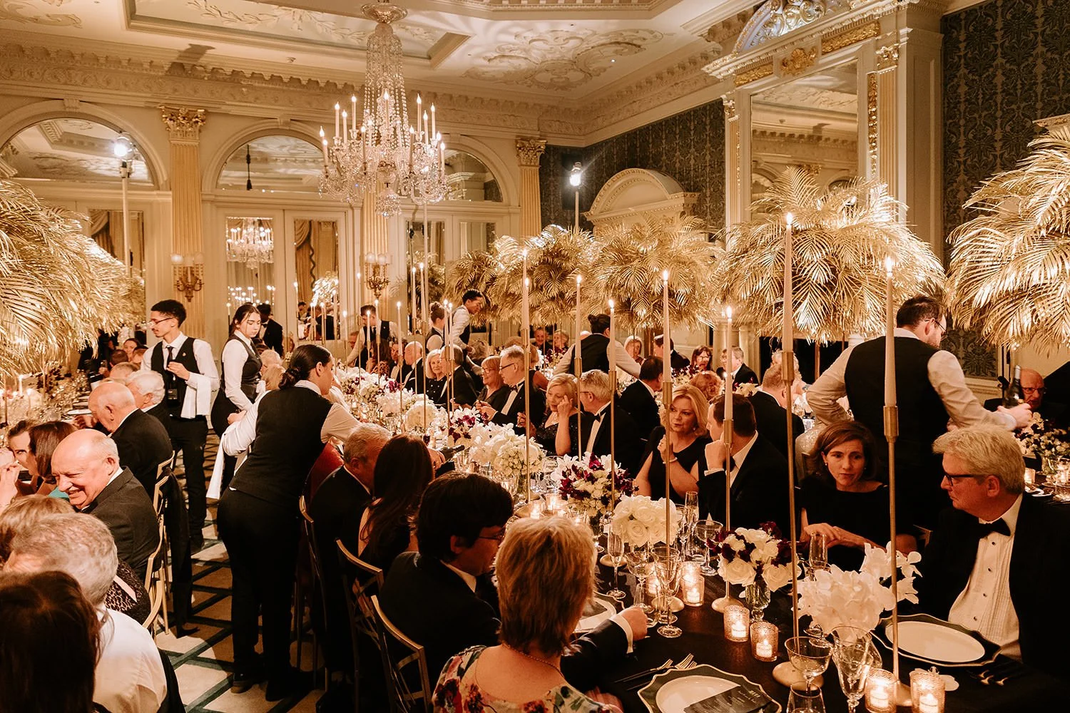 Elegant banquet hall with long dining table, floral centerpieces, candles, palm decorations, chandeliers, and well-dressed guests dining and socializing.