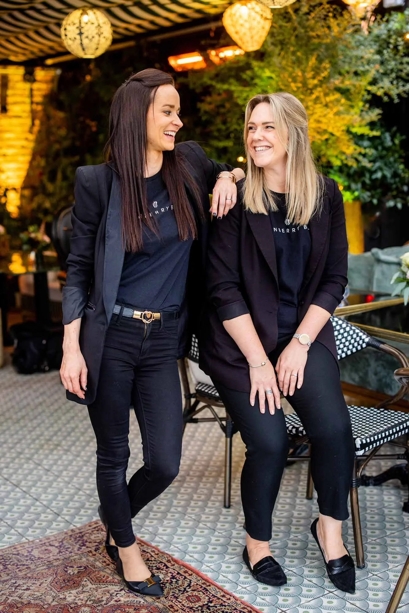 Two women dressed in black, standing and sitting in a decorated indoor space, smiling and enjoying each other's company.