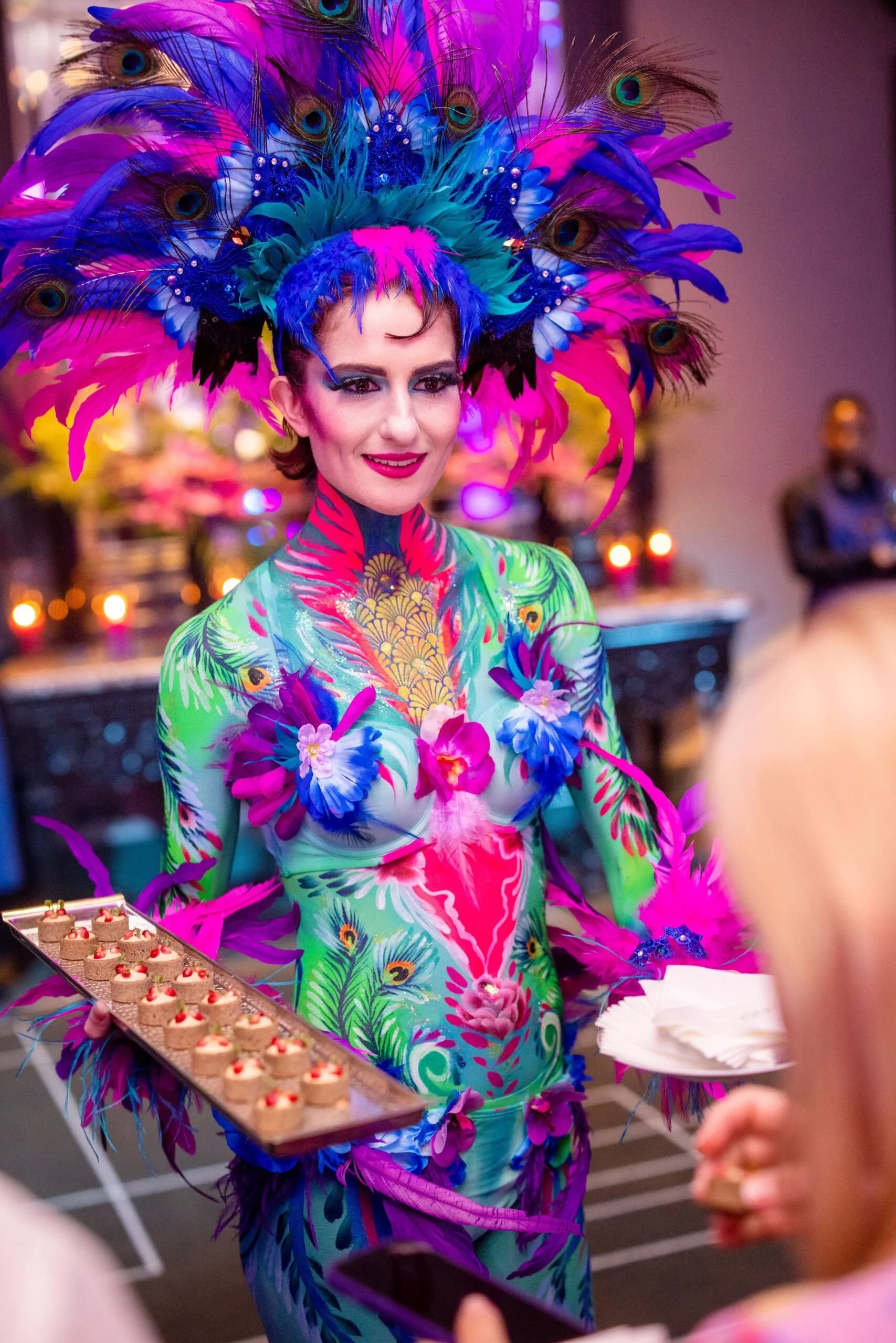A woman is dressed in a vibrant, colorful costume with a large feathered headdress featuring peacock feathers and bright pink, purple, and blue feathers. Her body is painted with elaborate, tropical-themed body art, including flowers and peacock feathers. She is smiling while holding a tray of small desserts.