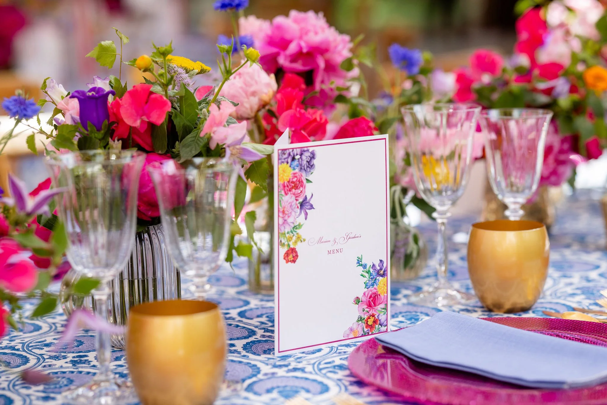 A table decorated with floral arrangements, wine glasses, gold cups, and a menu card with colorful flower illustrations.