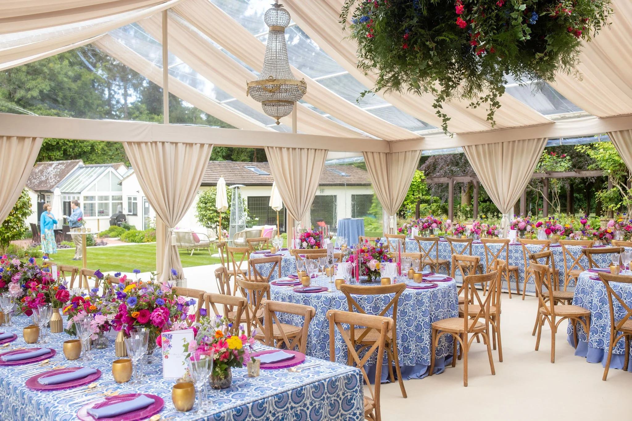 Elegant outdoor event tent decorated with floral arrangements, set for a celebration with multiple round tables, wooden chairs, and a chandelier hanging from the ceiling.