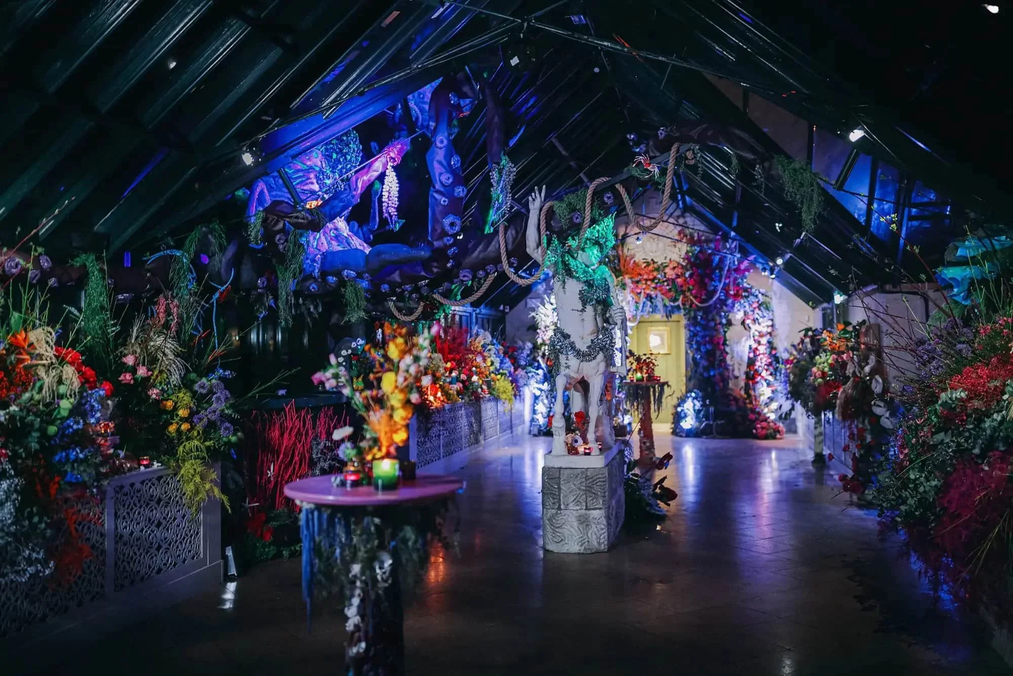 A dimly lit indoor space decorated with colorful flowers, plants, and hanging decorations, creating a vibrant and artistic atmosphere.