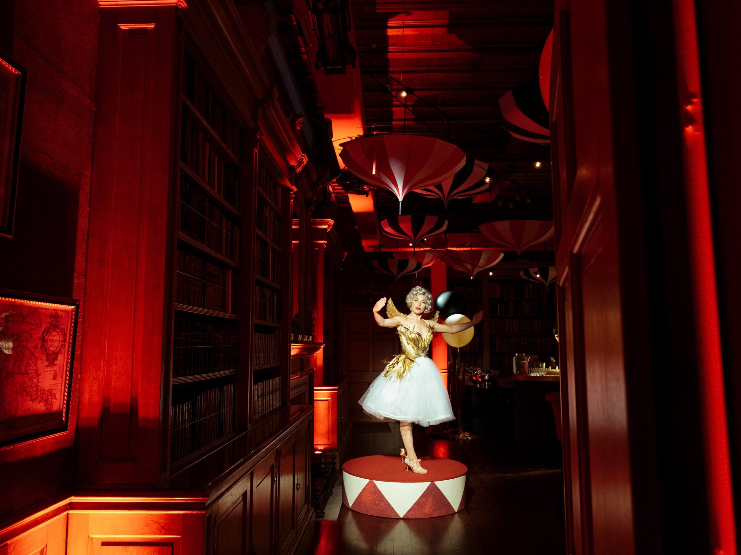A vintage mannequin dressed in a gold and white tutu with gold wings, holding balloons, standing on a red and white platform in a dimly lit room with dark wooden bookshelves and large pink and black umbrellas overhead.