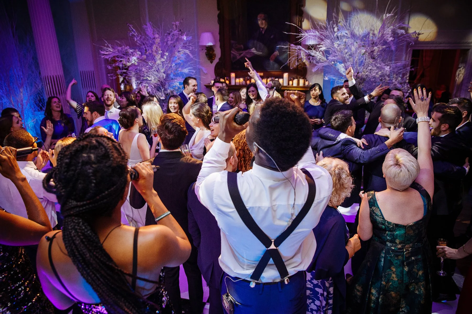 A lively indoor party with a large group of people dancing and celebrating, under purple and blue lighting, with decorative white floral arrangements and candles in the background.