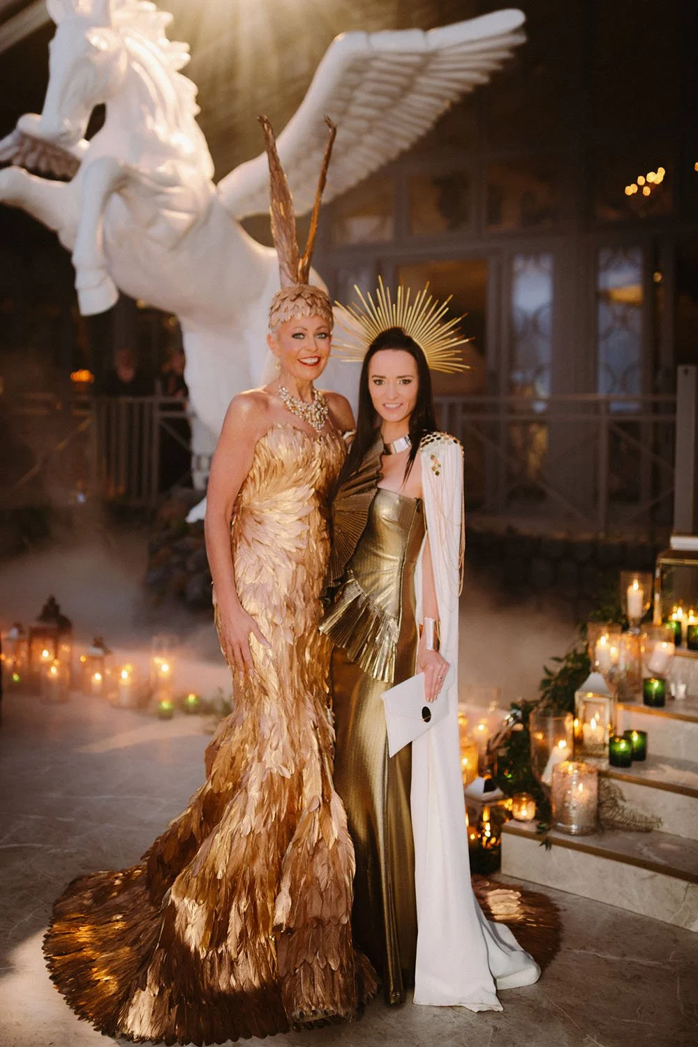 Two women in elaborate costumes standing together indoors, surrounded by candles and decorative lanterns, with a large white winged horse sculpture in the background.