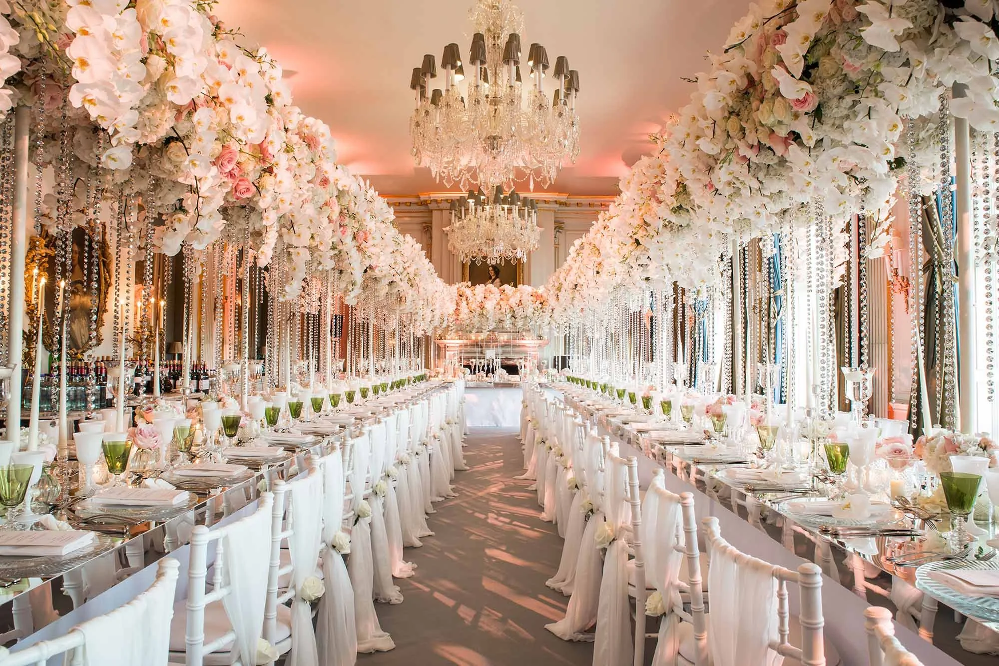 Elegant wedding reception decorated with white and pink flowers hanging from the ceiling, crystal chandeliers, and long banquet tables with white chairs, green glasses, and floral centerpieces.