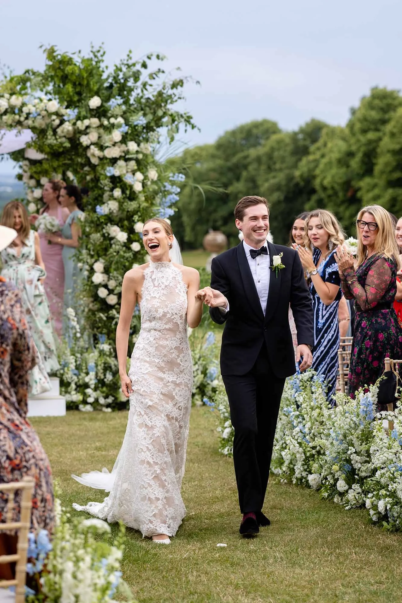 Bride and groom walking down the aisle after their wedding ceremony, smiling and holding hands. Guests are clapping and celebrating around them, with a floral arch and greenery in the background.