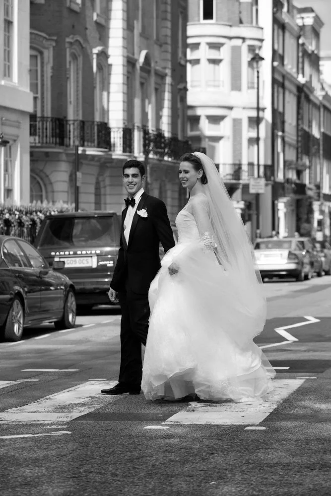 Black and white photo of a newlywed couple crossing the street, with the groom in a tuxedo and the bride in a wedding gown and veil, on a city street lined with parked cars and Victorian-style buildings.