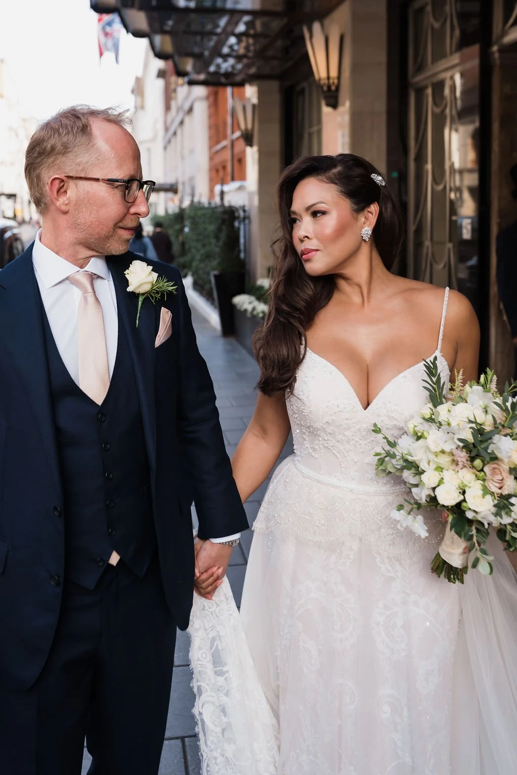 A bride in a white wedding dress holding a bouquet of white flowers and greenery, and a groom in a dark suit with a white shirt and tie, holding hands and looking at each other on a city street.