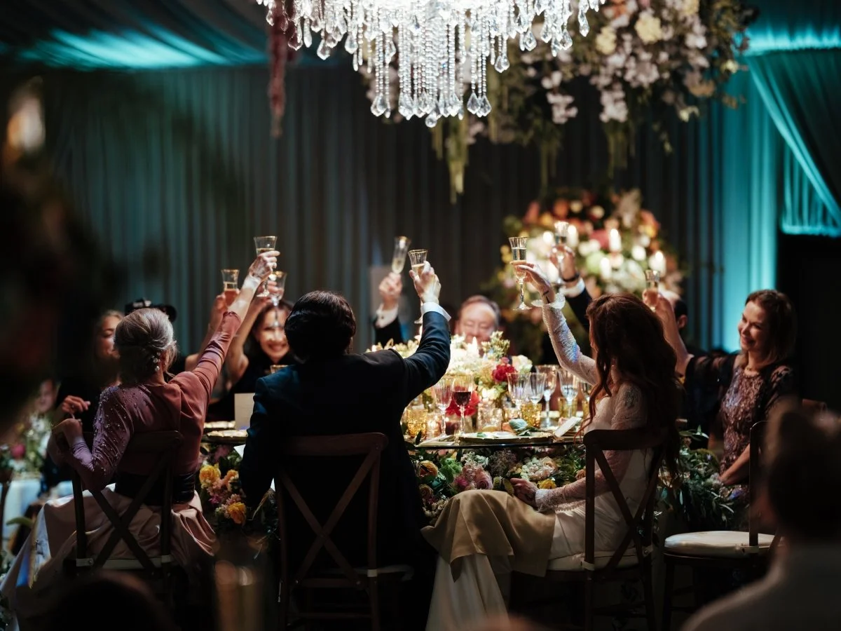 Guests at a wedding reception raising glasses in toast around a table with floral centerpieces and a chandelier overhead.