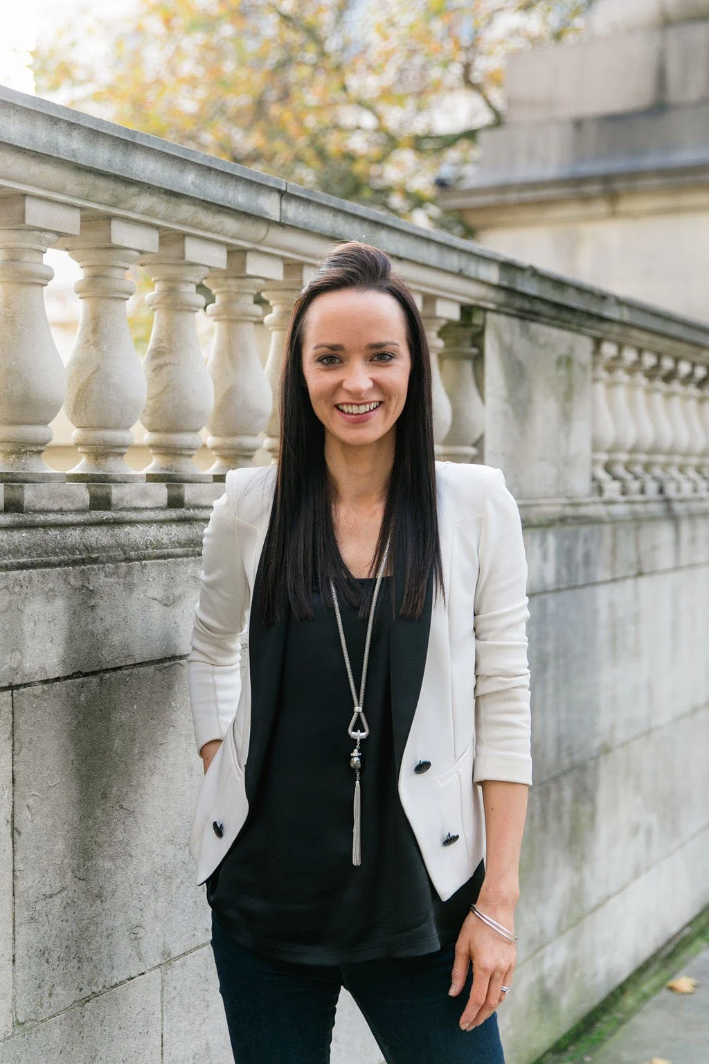 A woman with long dark hair, wearing a white blazer over a black shirt, standing outdoors in front of a stone balustrade, smiling at the camera.