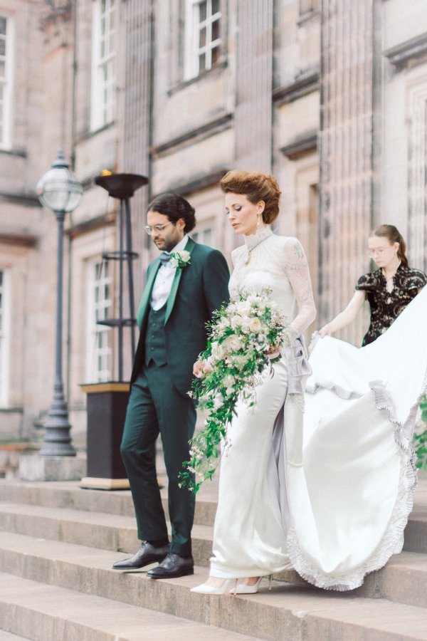 Bride and groom descend stone steps outside, bride holds bouquet and train, with attendant helping, in elegant wedding attire.