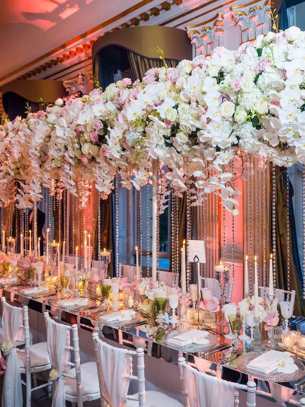 A lavish wedding banquet table with tall, cascading white and pink flowers and glowing candles in an elegant, ornate setting.