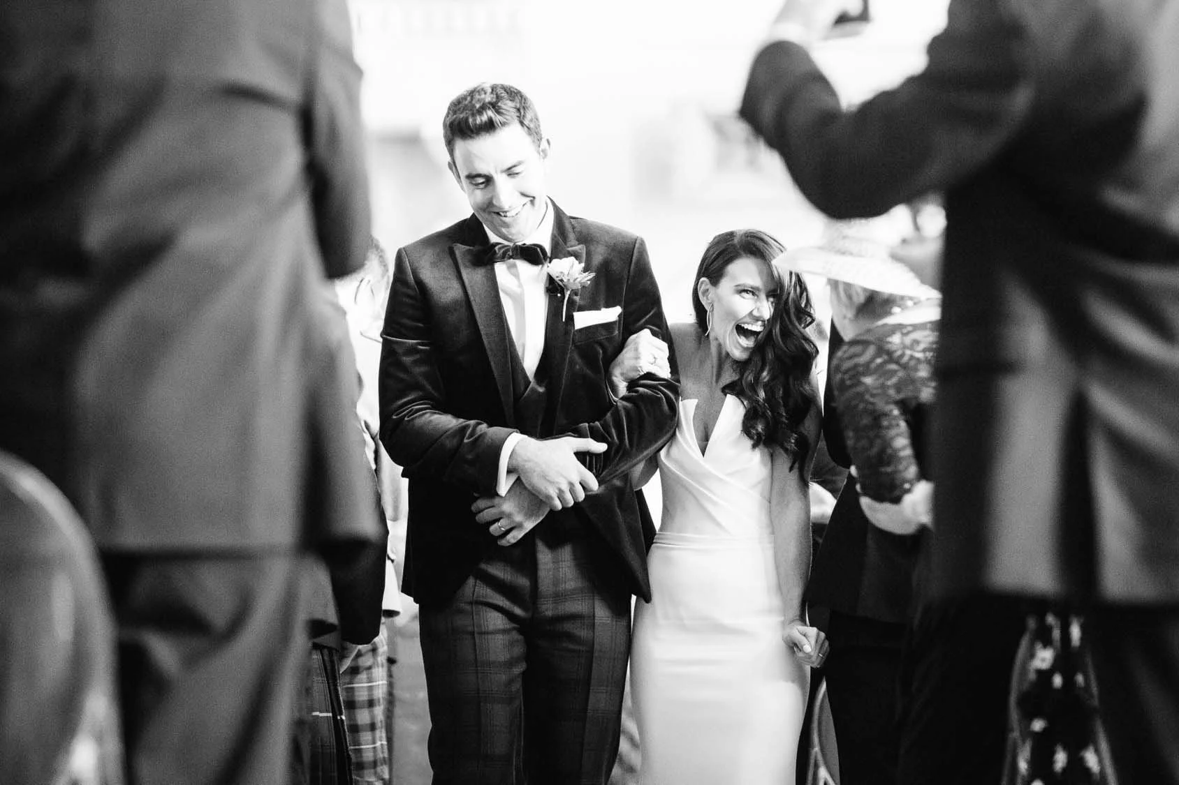 A joyful bride and groom walk arm in arm down the aisle, smiling and celebrating their wedding with guests around them.