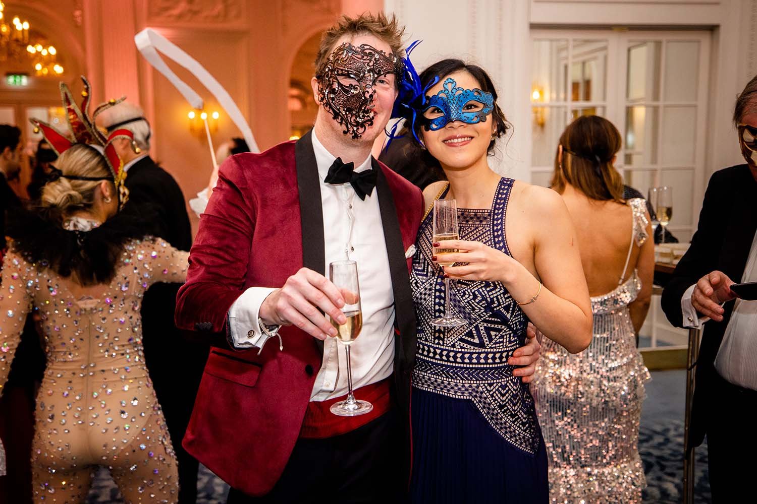 A man and woman in elegant attire celebrating at a masquerade party, both wearing decorative masks and holding glasses of champagne, with other party guests in costume in the background.