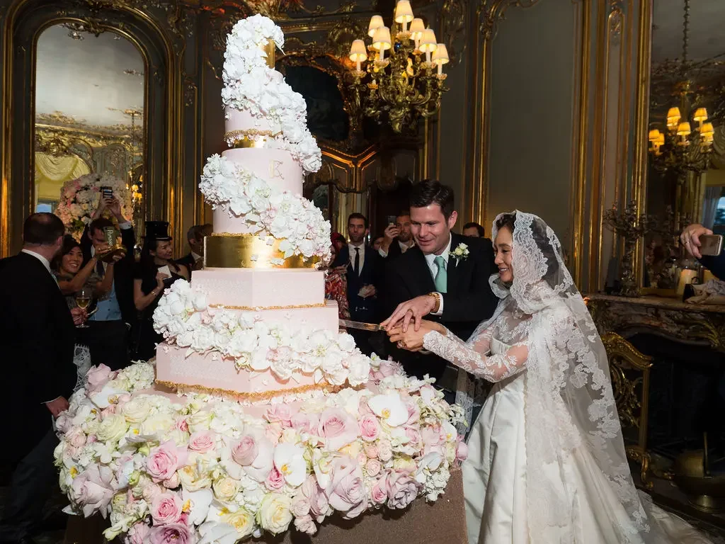 Bride and groom cutting a large, tiered wedding cake decorated with white flowers and gold accents, in an opulent room with gold mirrors, chandeliers, and ornate decor.