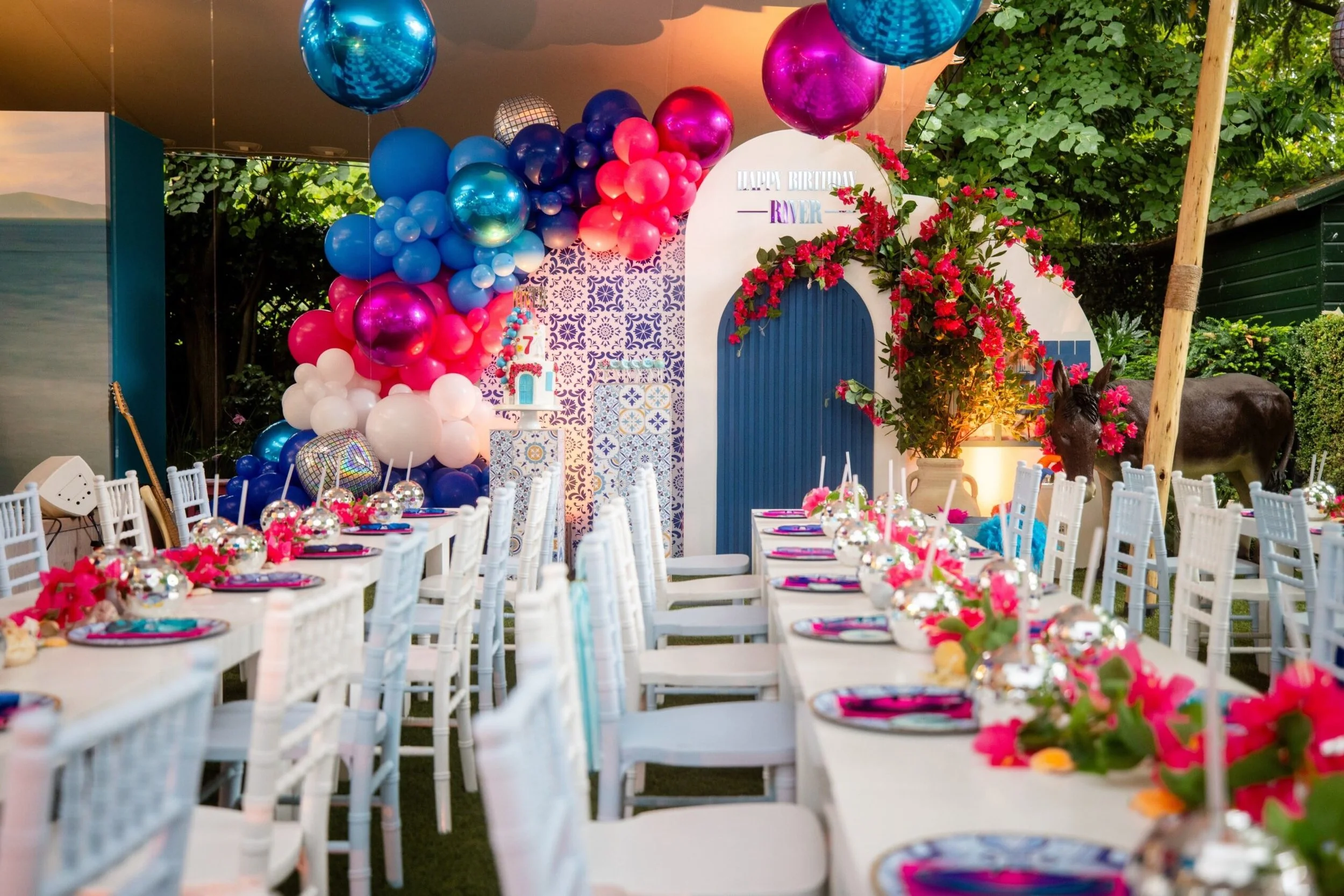 Decorated outdoor birthday party setup with white tables and chairs, pink, blue, and white balloons, pink flowers, and a blue door backdrop with a sign that reads 'Happy Birthday River'.