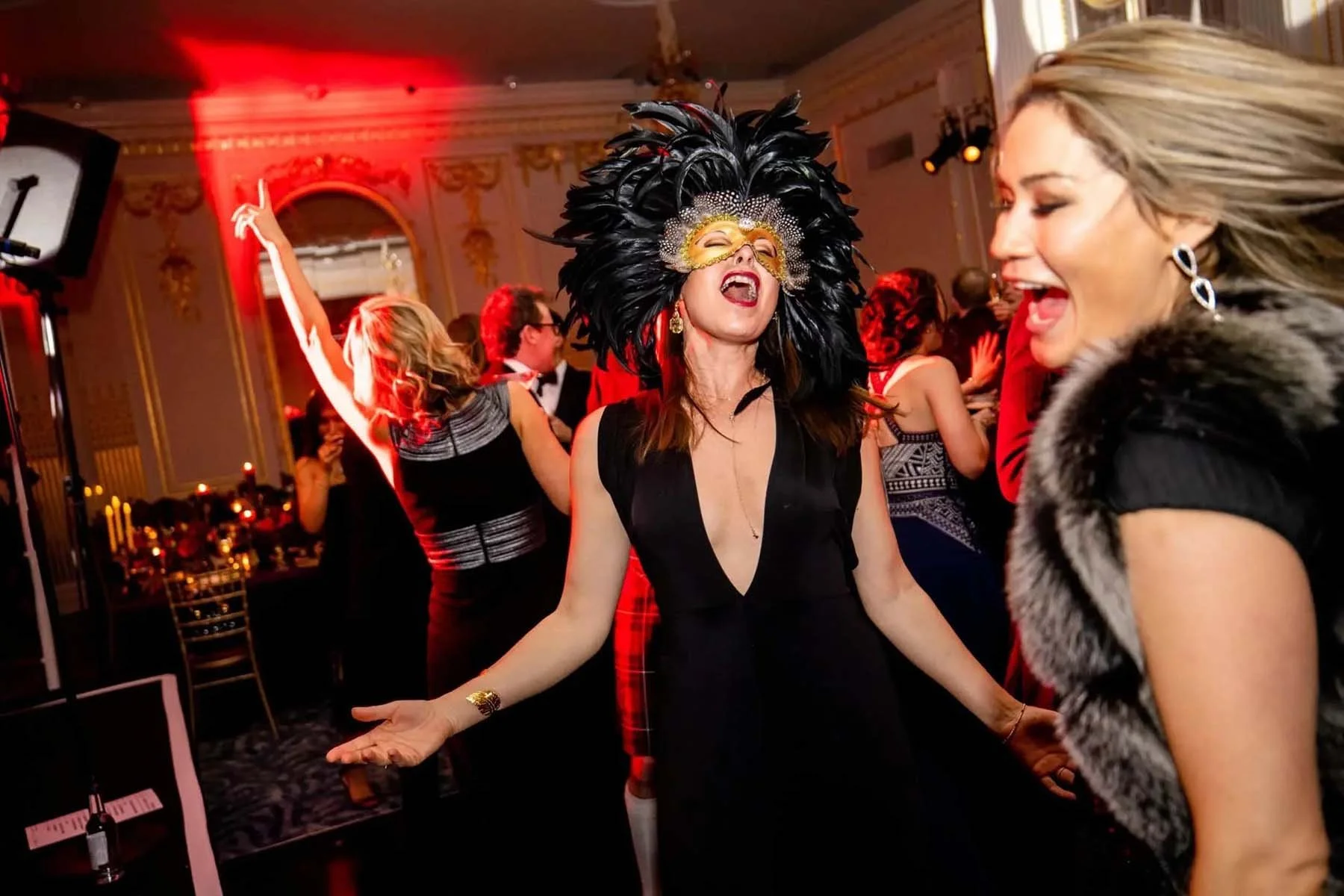 Women in elegant dresses, one wearing a feathered mask, joyfully dance and celebrate at a lively, upscale masquerade party.