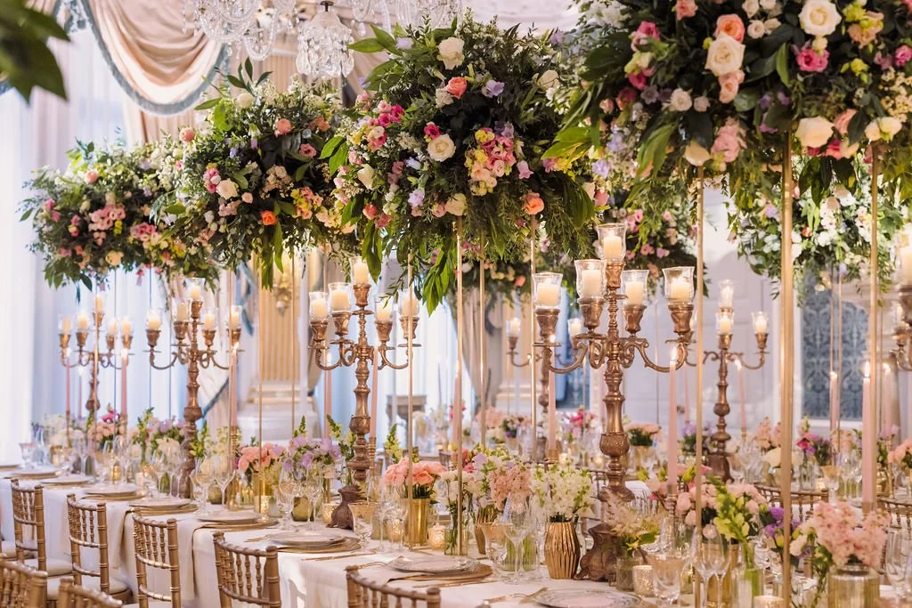 Elegant banquet table decorated with tall floral centerpieces, gold candelabras with lit candles, and floral arrangements in glass vases, set in a luxurious indoor venue with draped curtains and chandeliers.