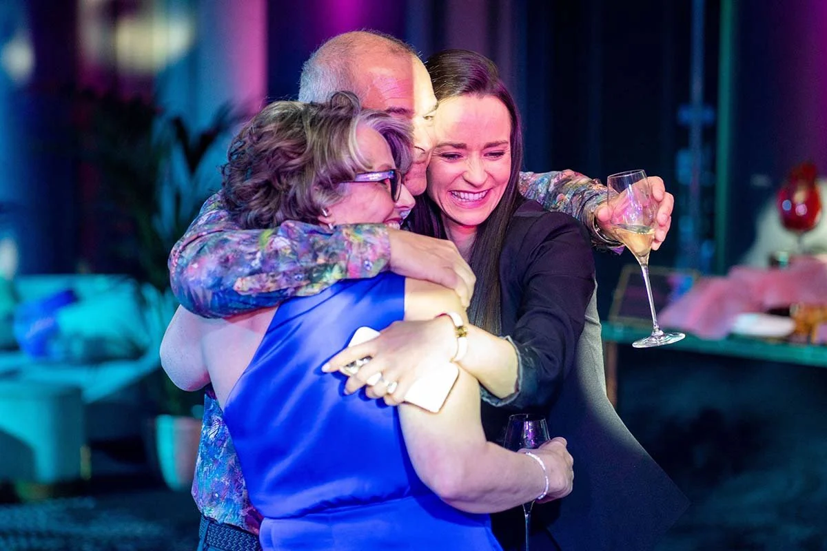 Three people celebrating, hugging, and holding glasses of champagne in a festive setting.