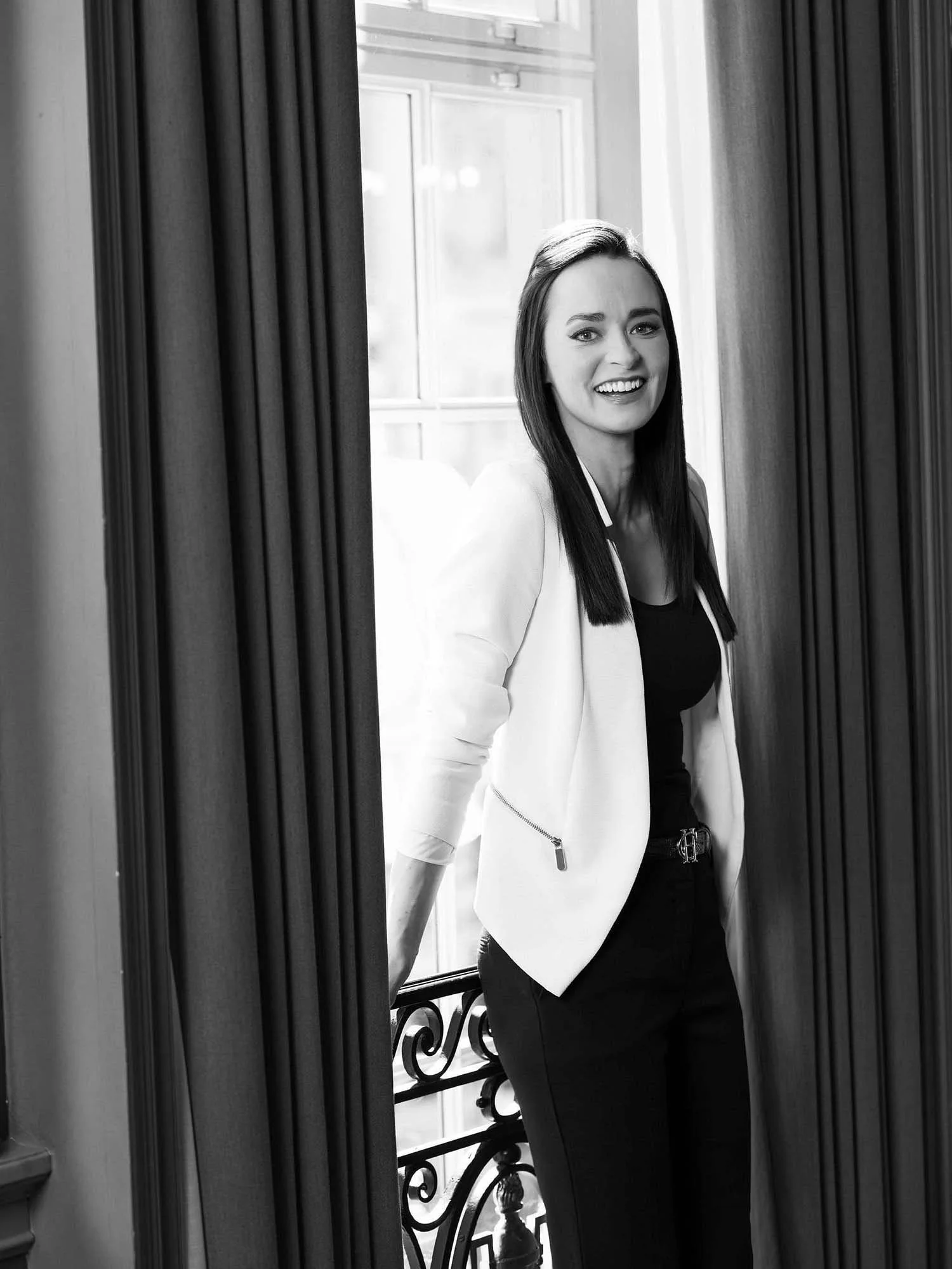Portrait of Melanie Helen in a white blazer smiles by a window between dark curtains in a black-and-white photo.