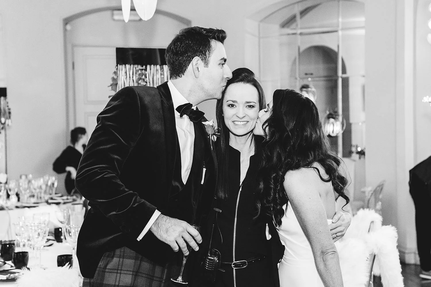 Black and white photo of a man in a tuxedo kissing a woman and a woman in a white dress kissing the man on the cheeks, smiling, at a formal gathering or celebration.