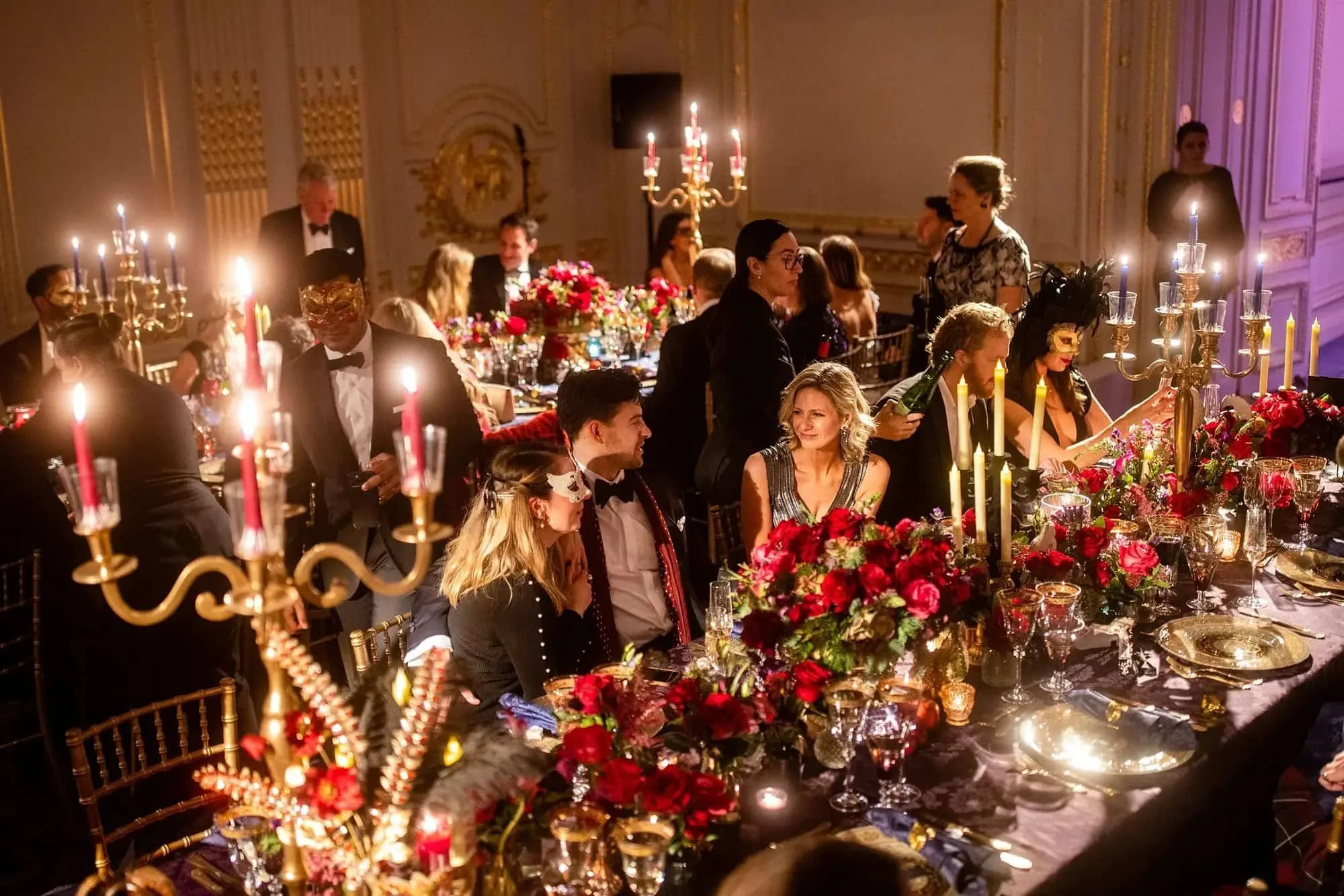 People in formal attire, some wearing masks, are gathered around a decorated banquet table with gold and red floral arrangements and candelabras, at an elegant event or party in a lavish, ornate room.
