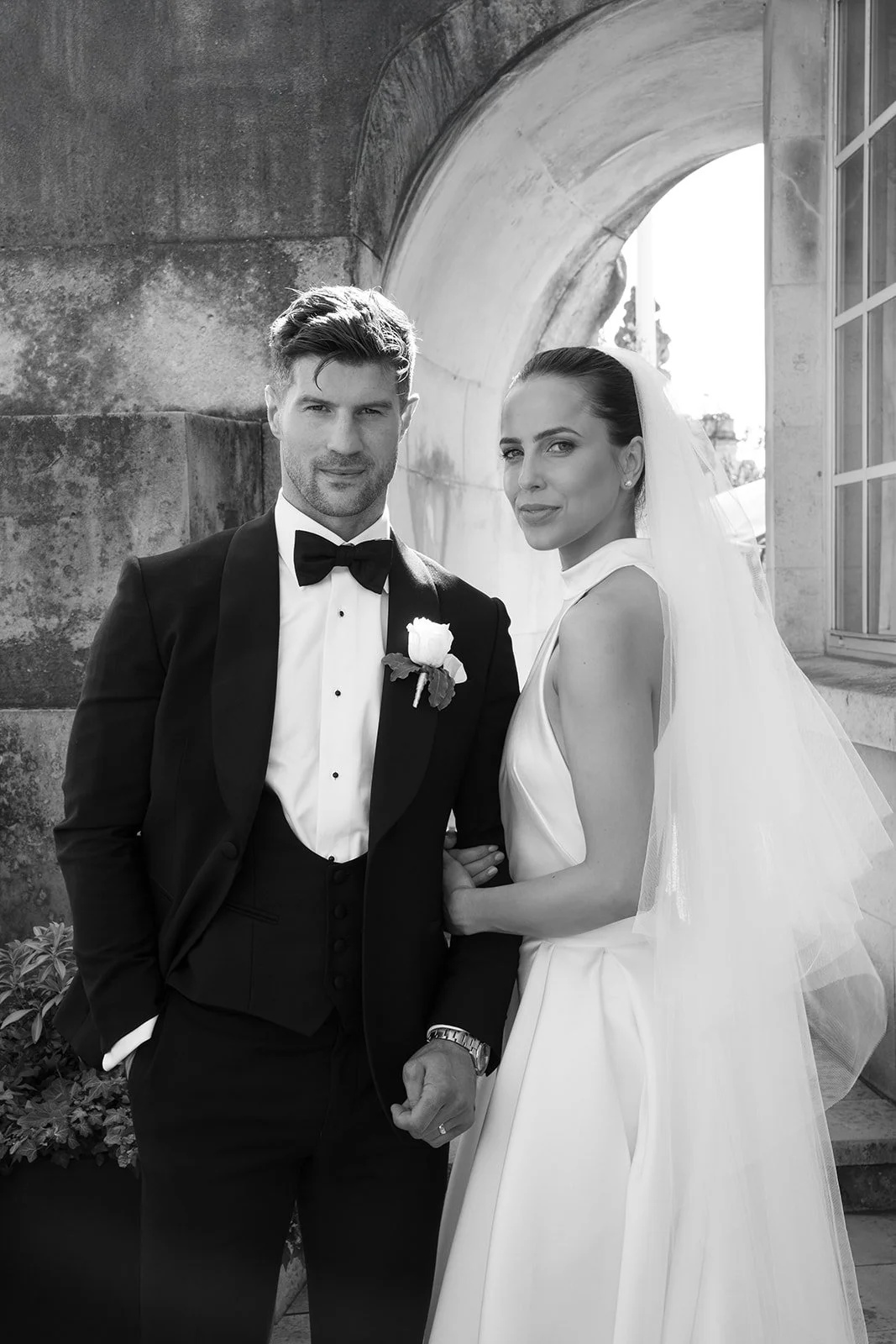 Black and white photo of a bride and groom standing outside near an arched stone structure. The groom is wearing a tuxedo with a bow tie and a white rose boutonniere, while the bride is wearing a sleeveless wedding gown with a veil.