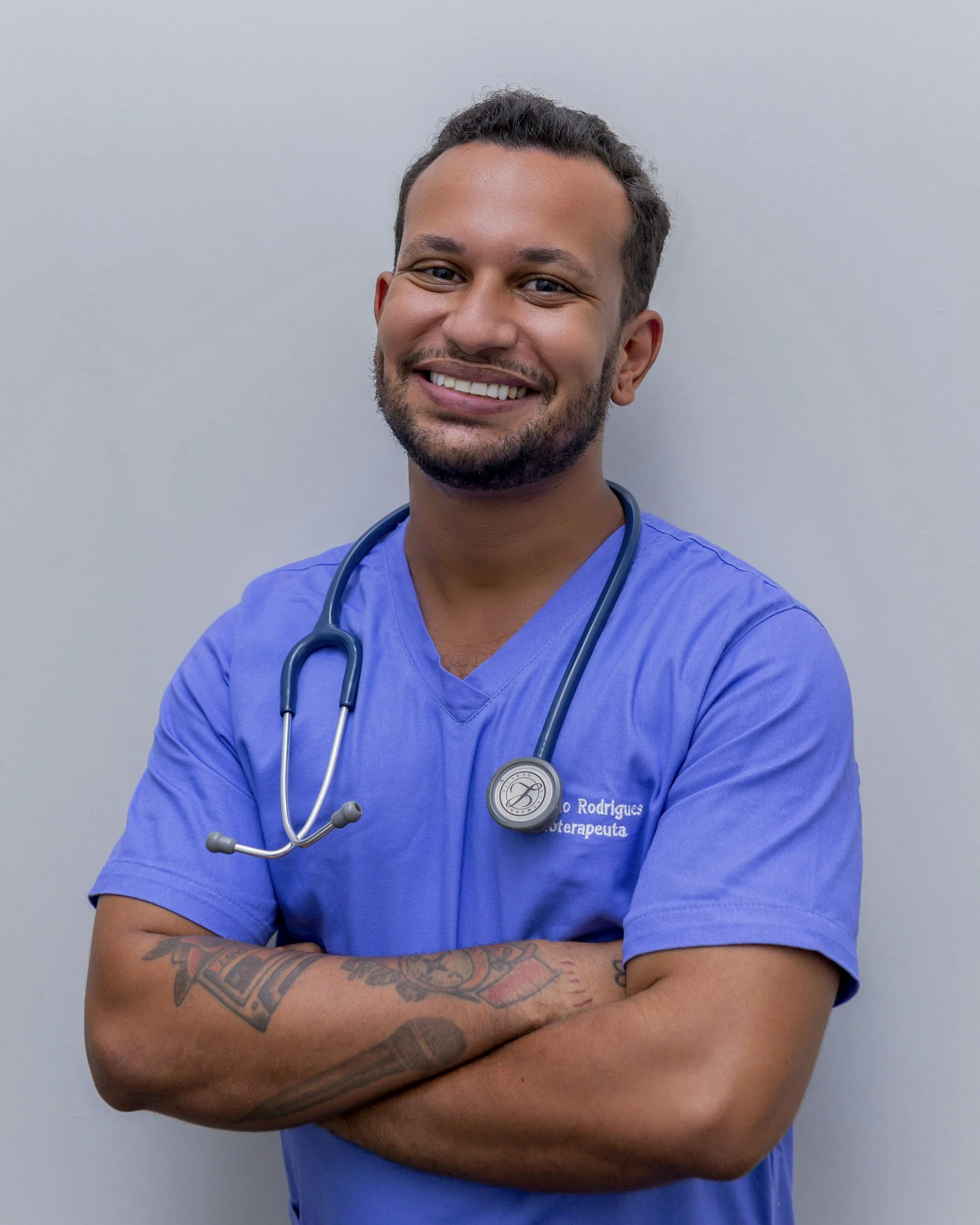 A smiling male healthcare professional wearing blue scrubs and a stethoscope around his neck, standing with arms crossed against a plain grey background.