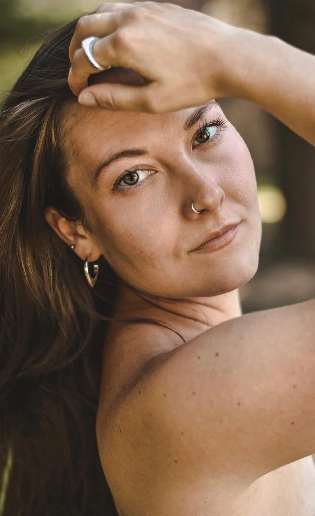 Close-up of a woman with long brown hair, wearing earrings and a nose ring, resting her hand on her forehead. She has a neutral expression and is looking at the camera outdoors.