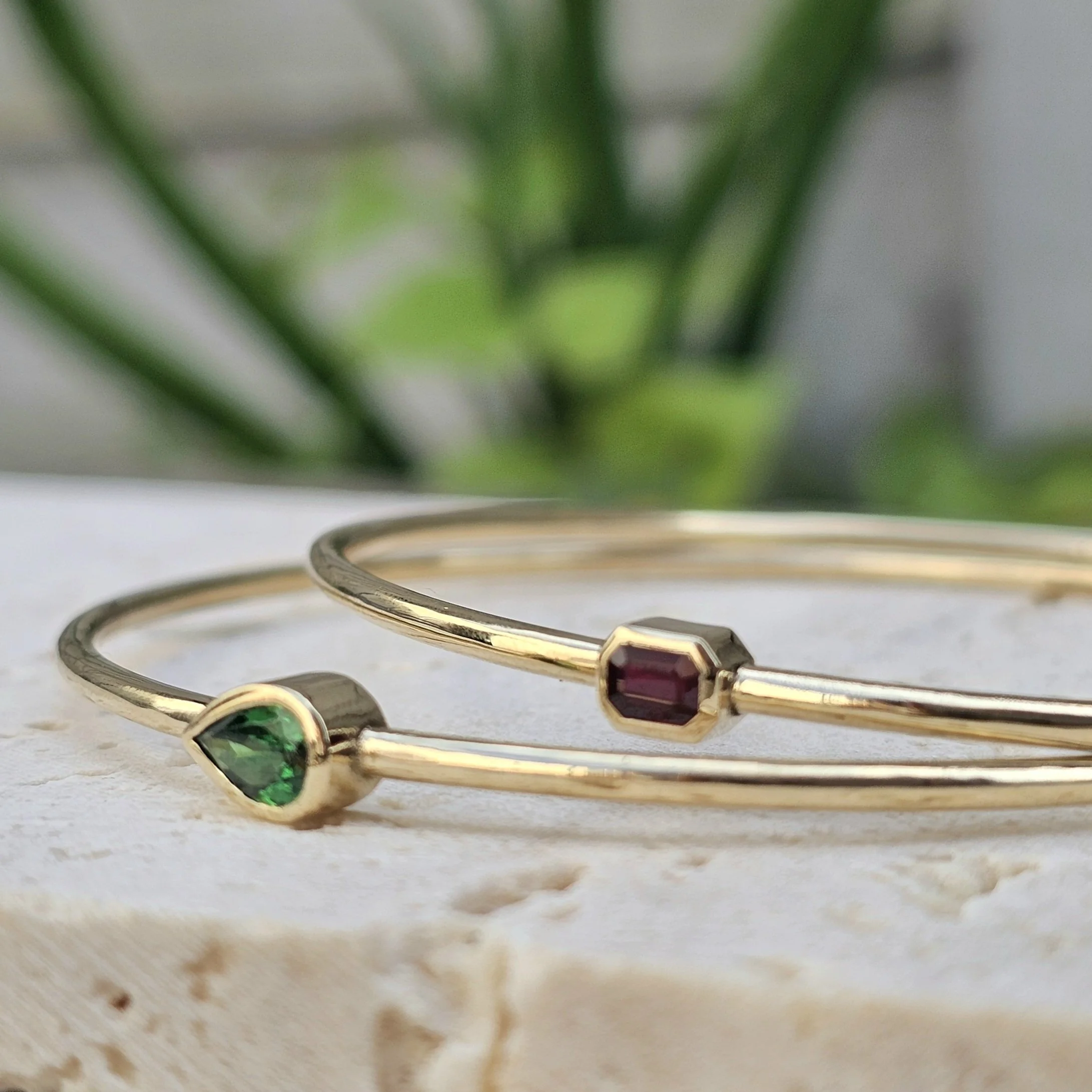 Close-up of a yellow bracelet with green, red, and purple gemstones on a white surface, with blurred green foliage in the background.