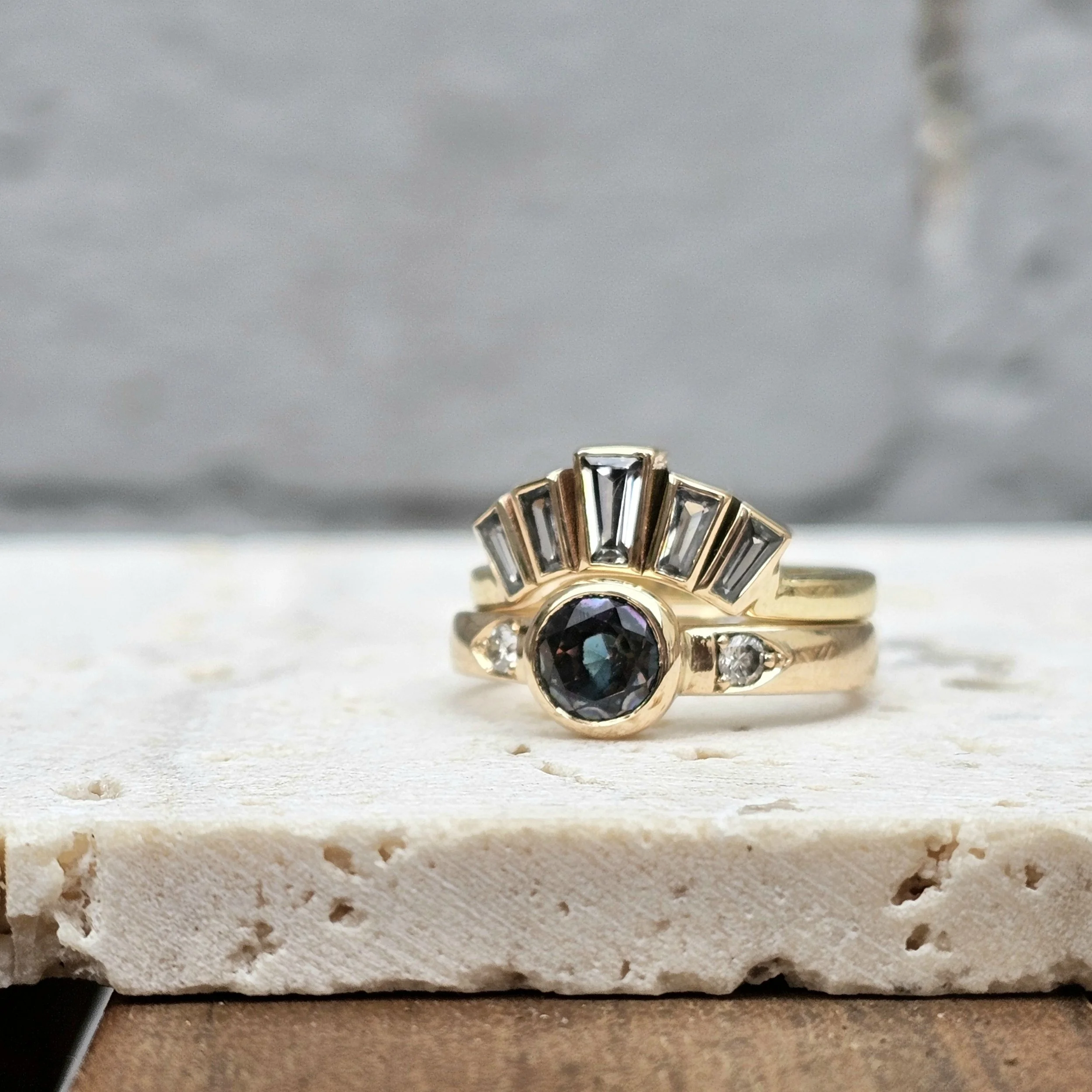 A close-up of a gold ring with a blue topaz central gemstone, layered with other rings featuring small gemstones, resting on a textured white surface.