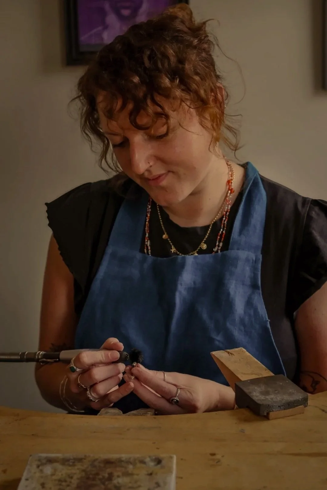 A woman with curly red hair wearing a dark shirt, multiple necklaces, and a blue apron, working with jewelry tools at a wooden workbench.