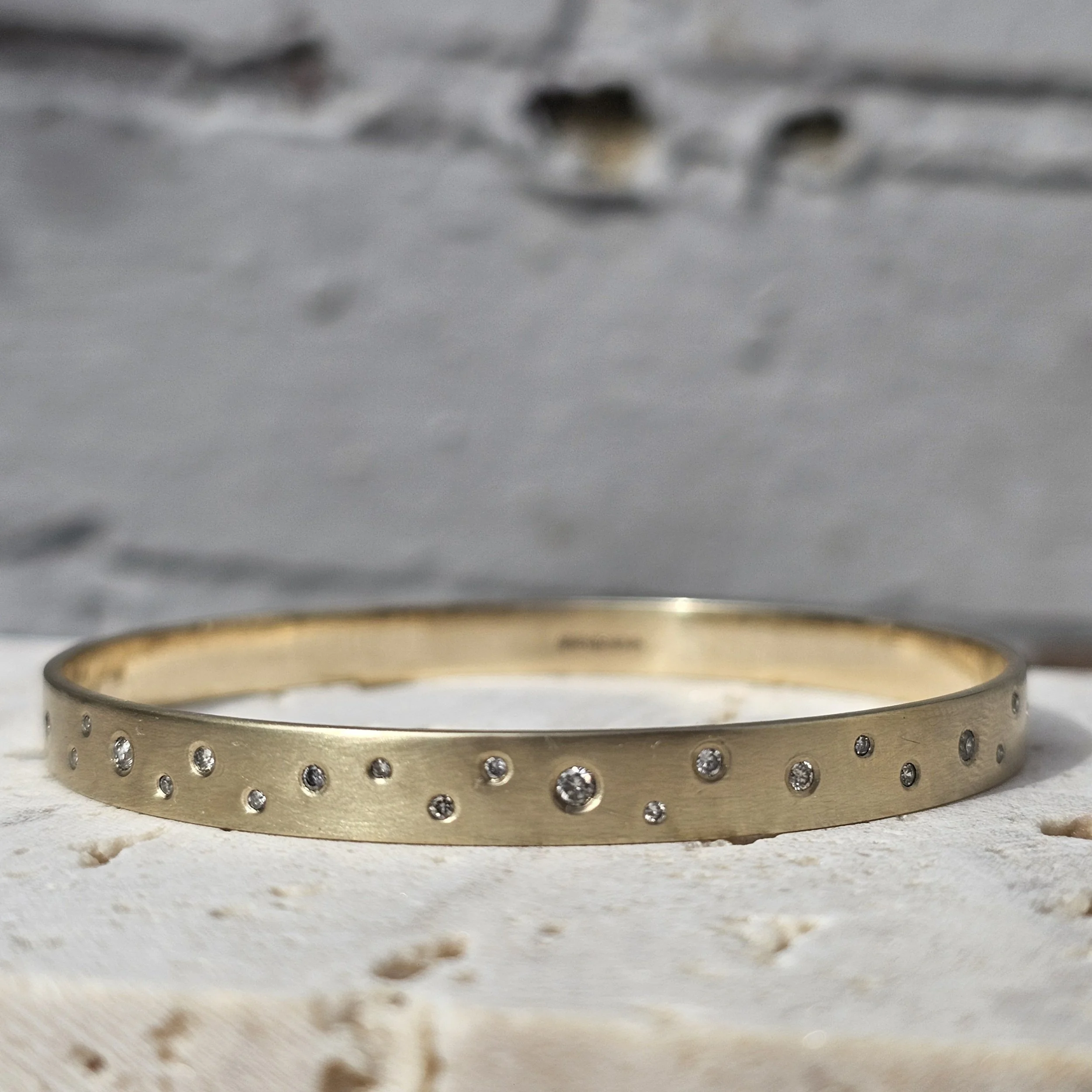 close-up of a gold bangle with diamonds set in a scattered fashion, resting on a textured surface with a grey background.