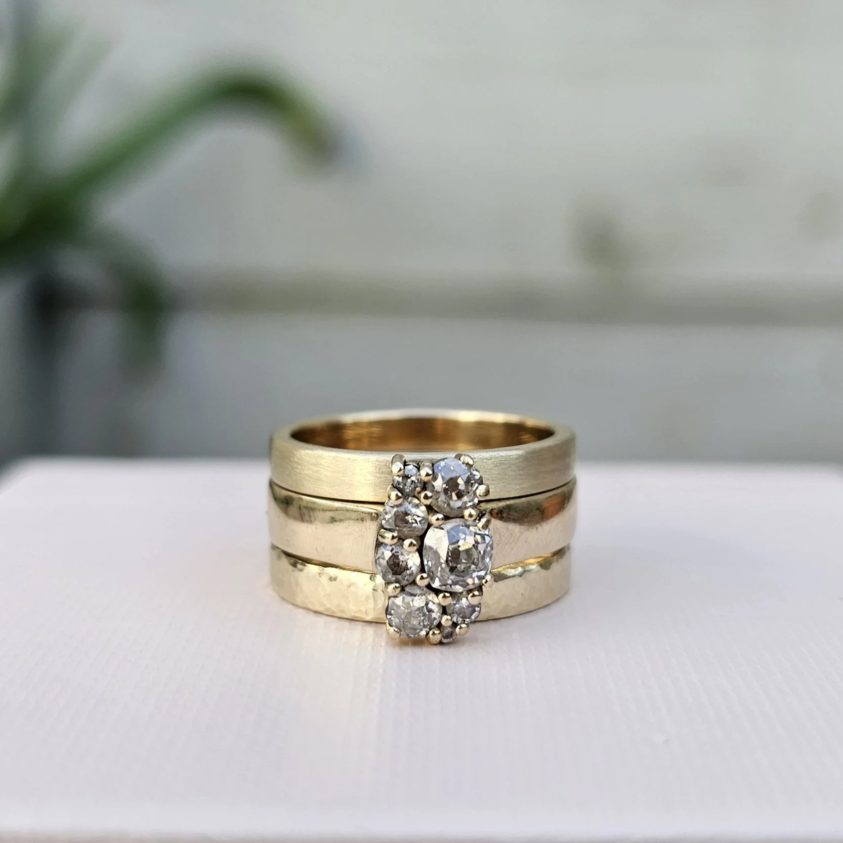Close-up of stacked gold rings, one with a cluster of diamonds, on a white surface with a blurred green plant in the background.