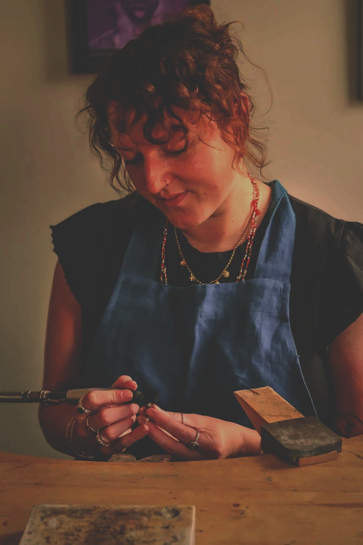 A woman with curly hair, wearing a blue apron and multiple necklaces, is working with tools at a wooden workbench in a dimly lit room.