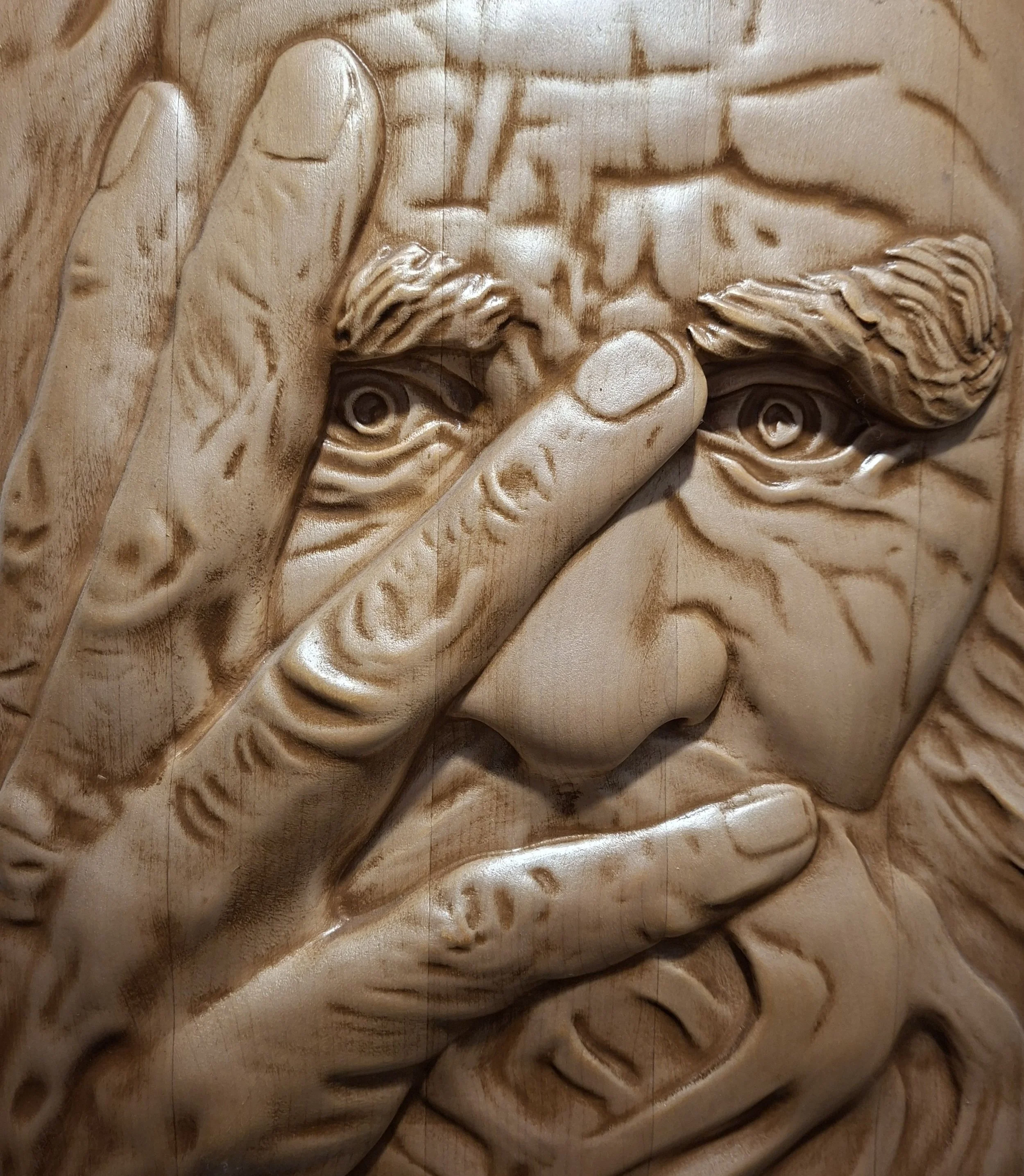 Close-up of a wood relief sculpture showing a man's face with detailed carved features, including a full beard and a hand raised to his forehead.