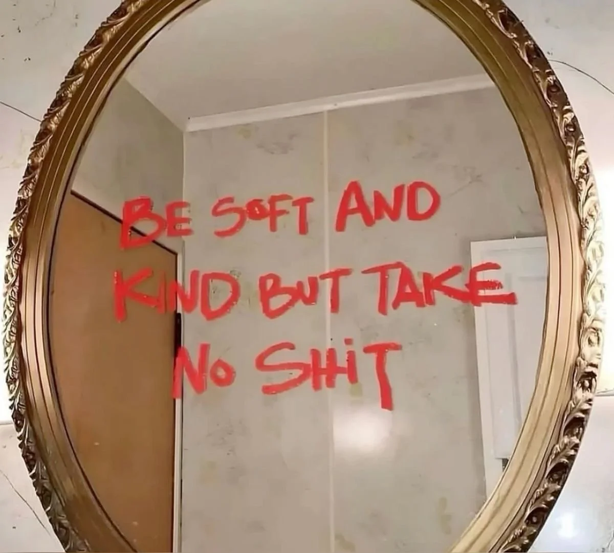 A mirror with a decorative gold frame displaying a humorous message written in red ink, which says: 'Be soft and kind but take no shit'. The reflection shows part of a wall, a ceiling, and a door in a room.