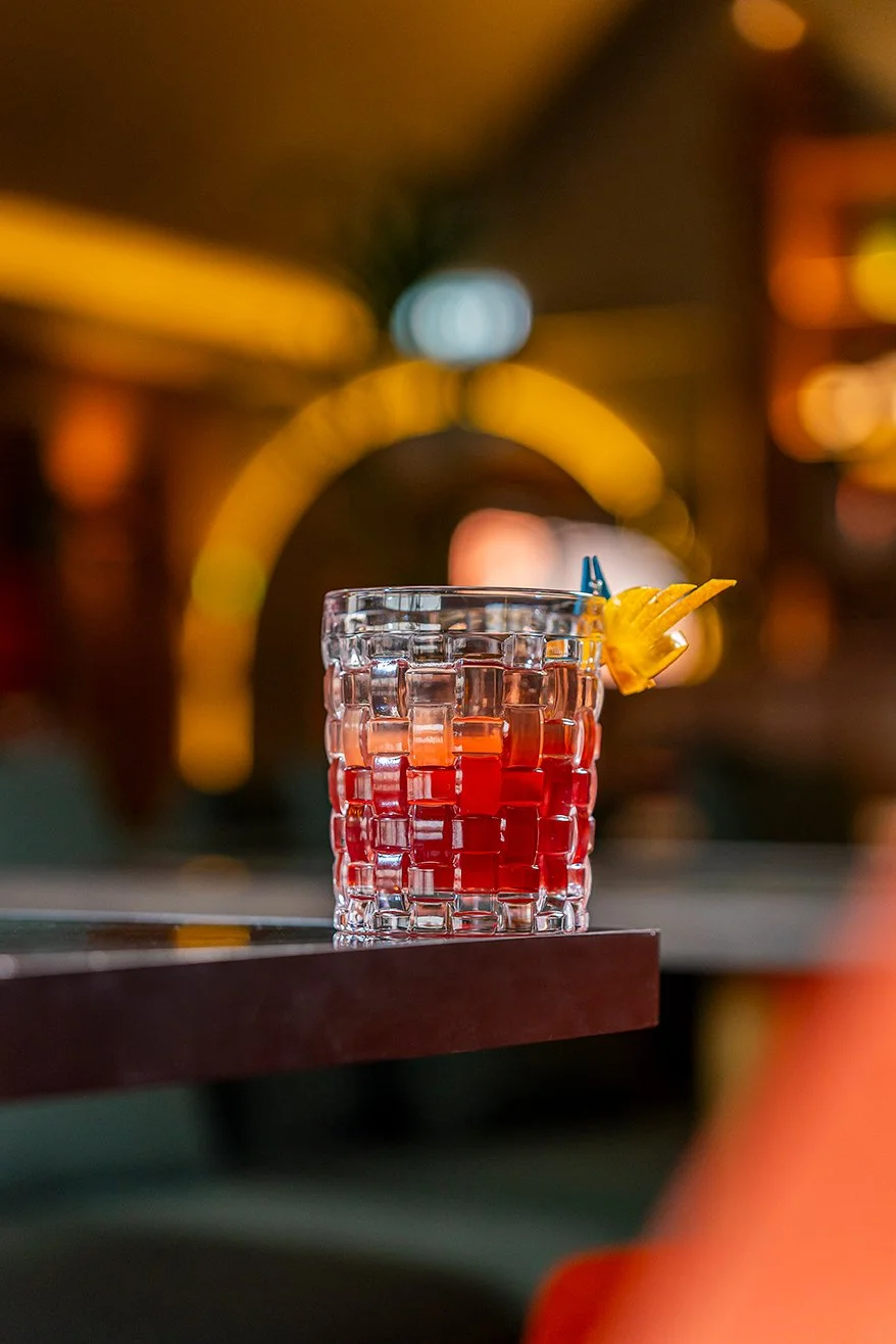 Copo de coquetel com líquido vermelho e gelo, decorado com uma fatia de abacaxi e um canudo azul, com iluminação ambiente difusa ao fundo.