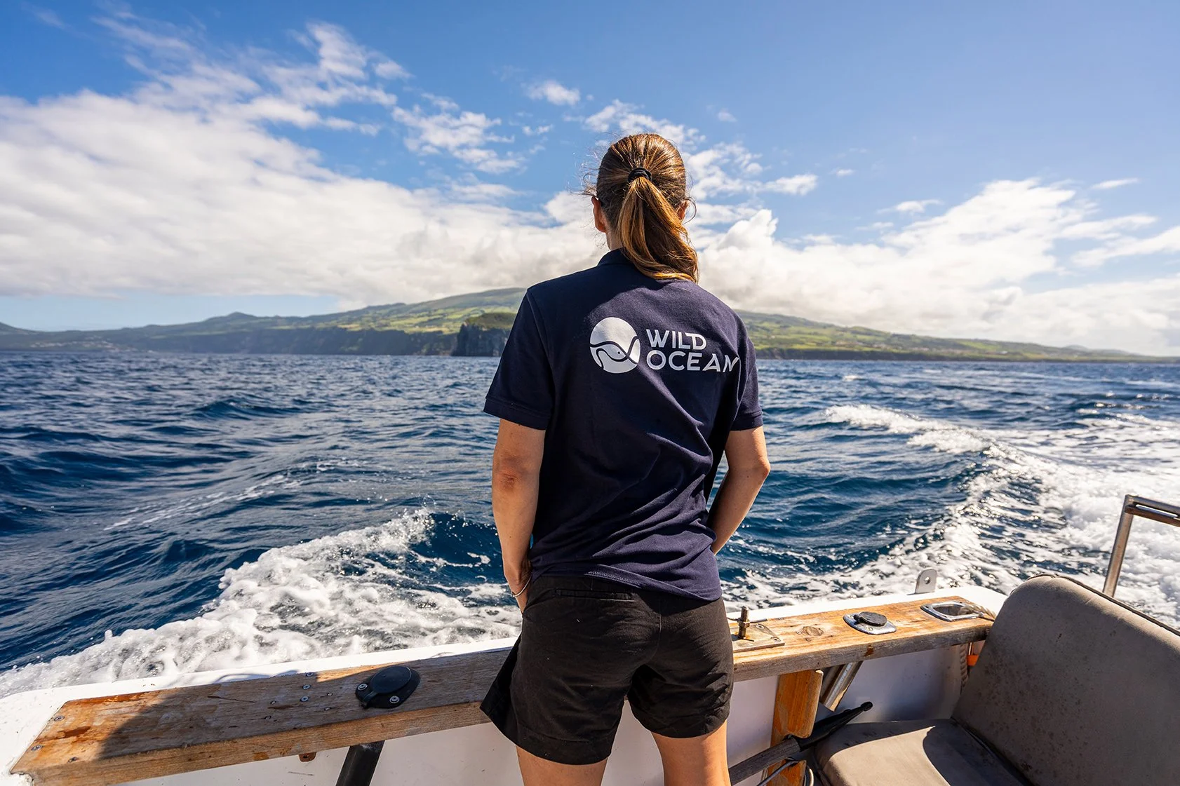 Mulher de costas, usando camiseta preta com logo 'Wild Ocean', observando o mar aberto em um barco, com ilha ao fundo sob céu parcialmente nublado.