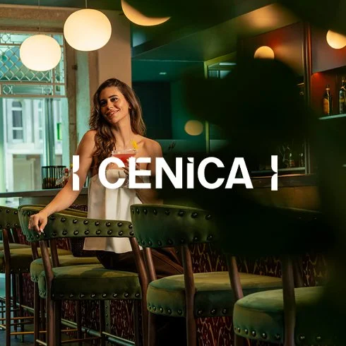 Mulher sorridente sentada em um bar ou restaurante moderno, com cadeiras verdes, luzes suspensas de forma de globos e decoração contemporânea.