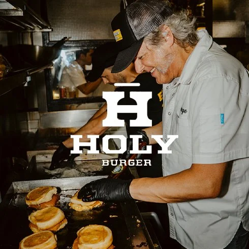 Homem idoso sorridente, vestindo uniforme e máscara, preparando hambúrgueres na cozinha do restaurante Holy Burger.