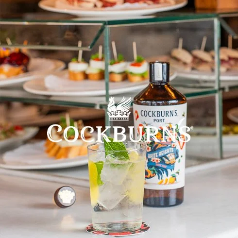 Copo com drink, garrafinha de gin Cockburns e petiscos em uma bandeja.