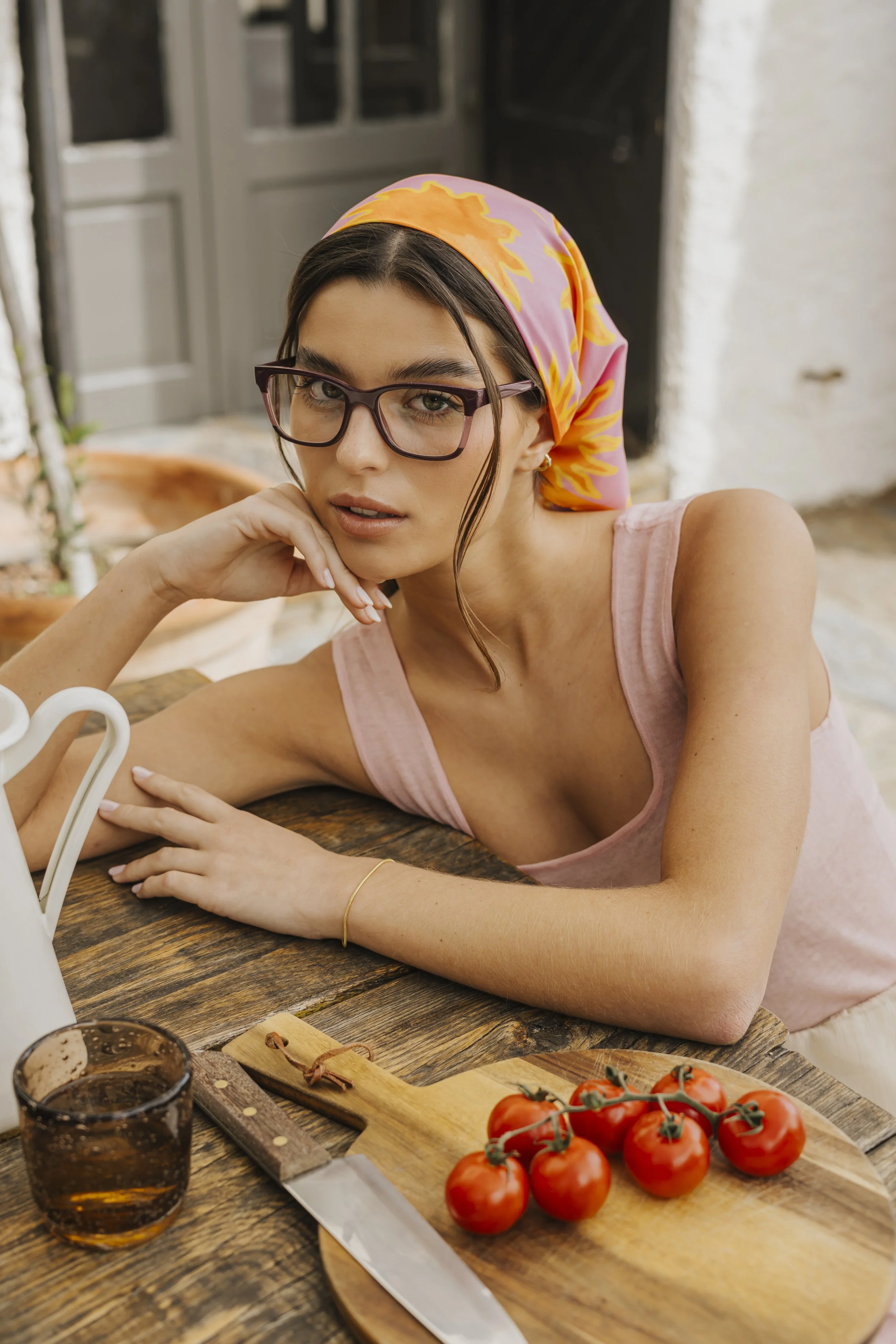 Mulher com óculos e lenço colorido na cabeça, deitada sobre uma mesa de madeira, ao lado de tomates e uma faca, num ambiente externo.