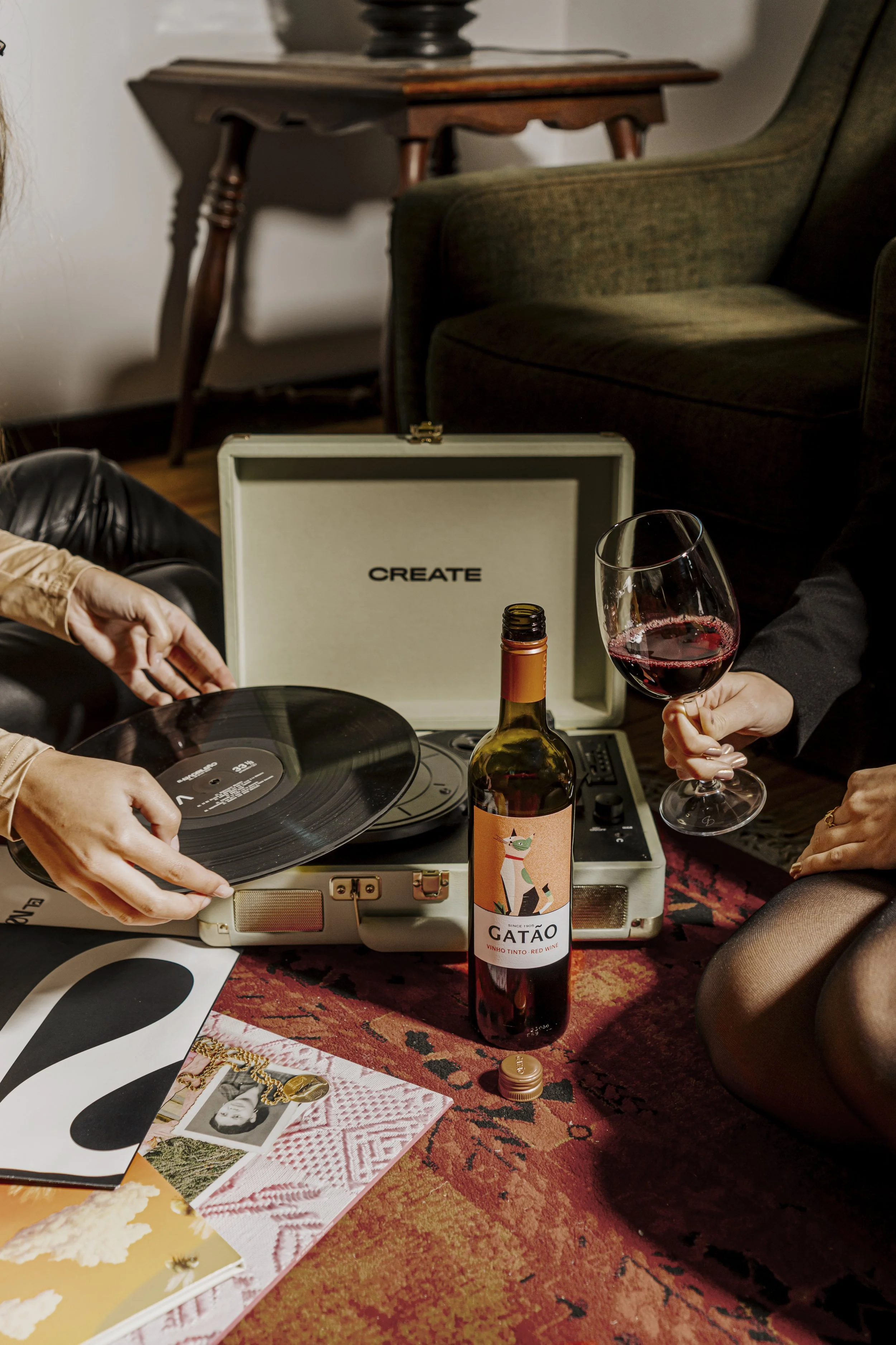 Uma festa com um toca-discos vintage ao centro, tocando um disco de vinil, com uma pessoa de mão ajudando a colocar o disco, ao lado uma garrafa de vinho tinto com rótulo que diz 'Gatão', uma taça de vinho tinto na mão de uma pessoa, vários discos de vinil e revistas ao redor, em um ambiente aconchegante.