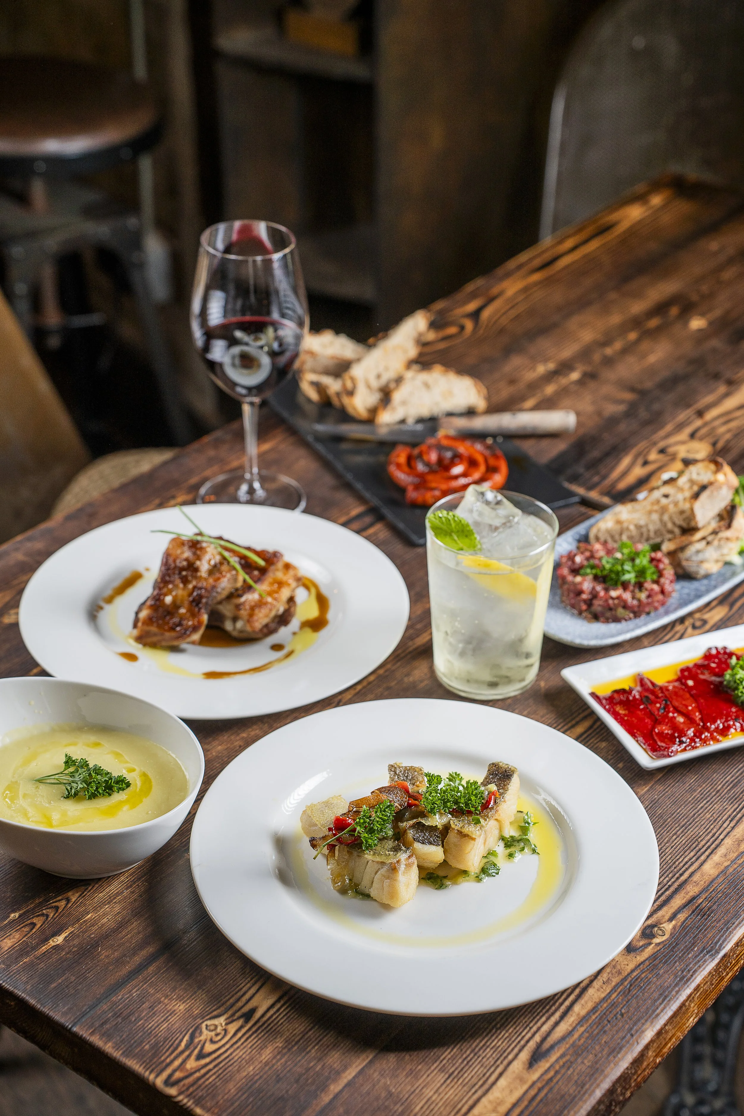 Mesa de jantar com vários pratos de comida, um copo de drink com gelo, e uma taça de vinho tinto.