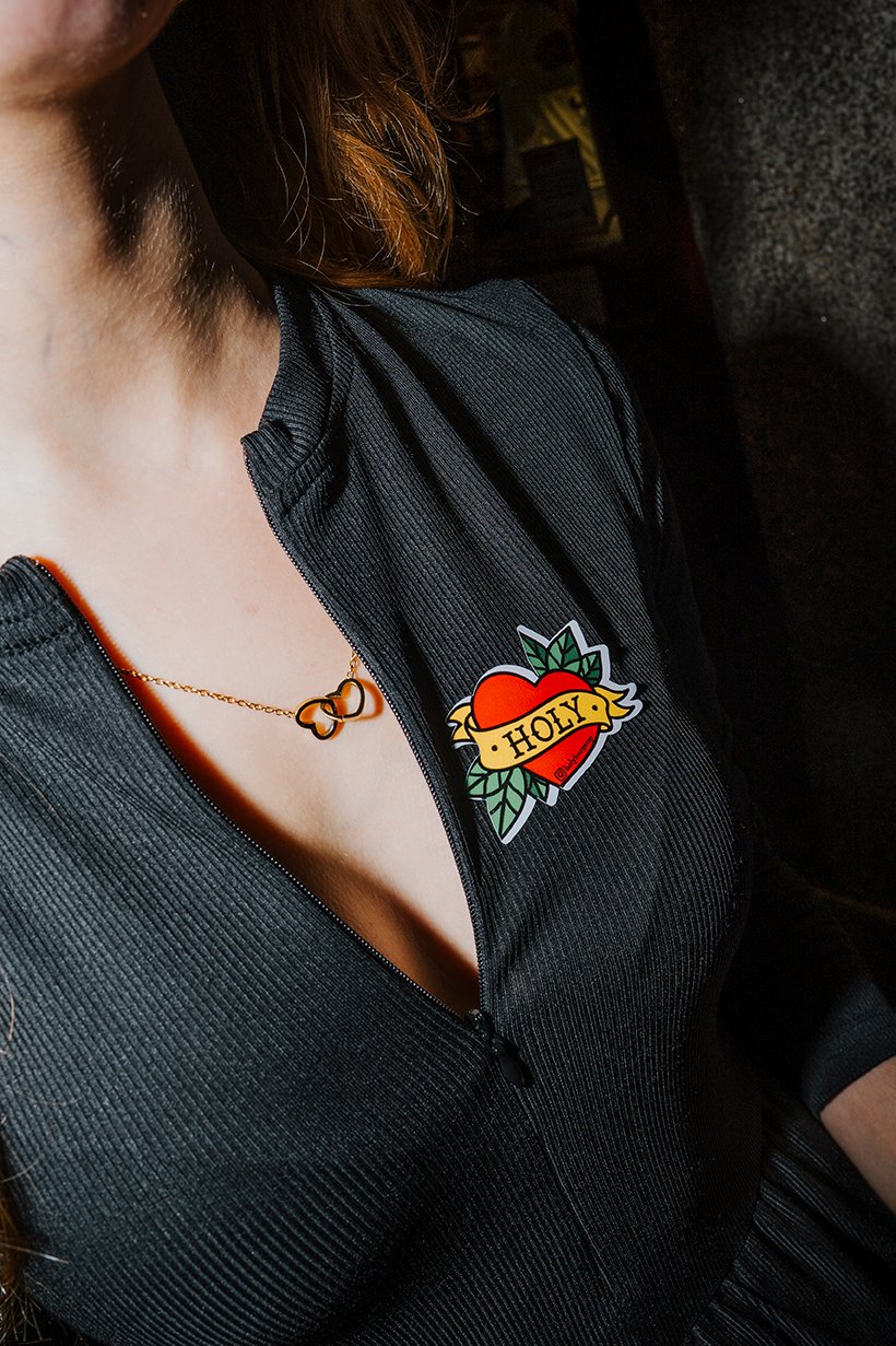 Close-up de uma mulher usando uma jaqueta preta com um broche de coração com a palavra 'Holy' e uma corrente dourada com um pingente de laço.