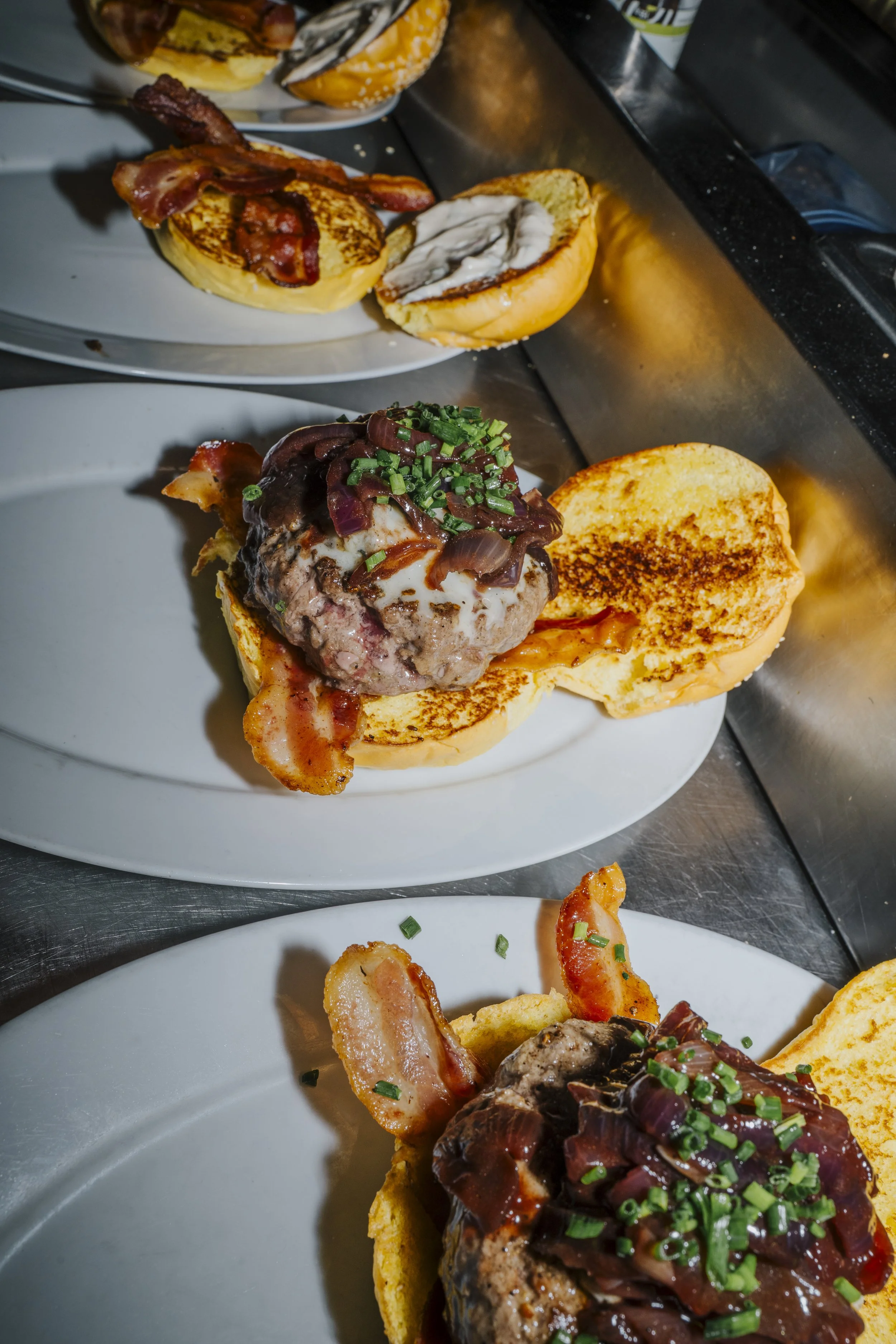 Três pratos de hambúrguer com diferentes carnes, bacon, queijo, cebola e chapeados, servidos em pães tostados, sobre pratos brancos em uma bancada de cozinha